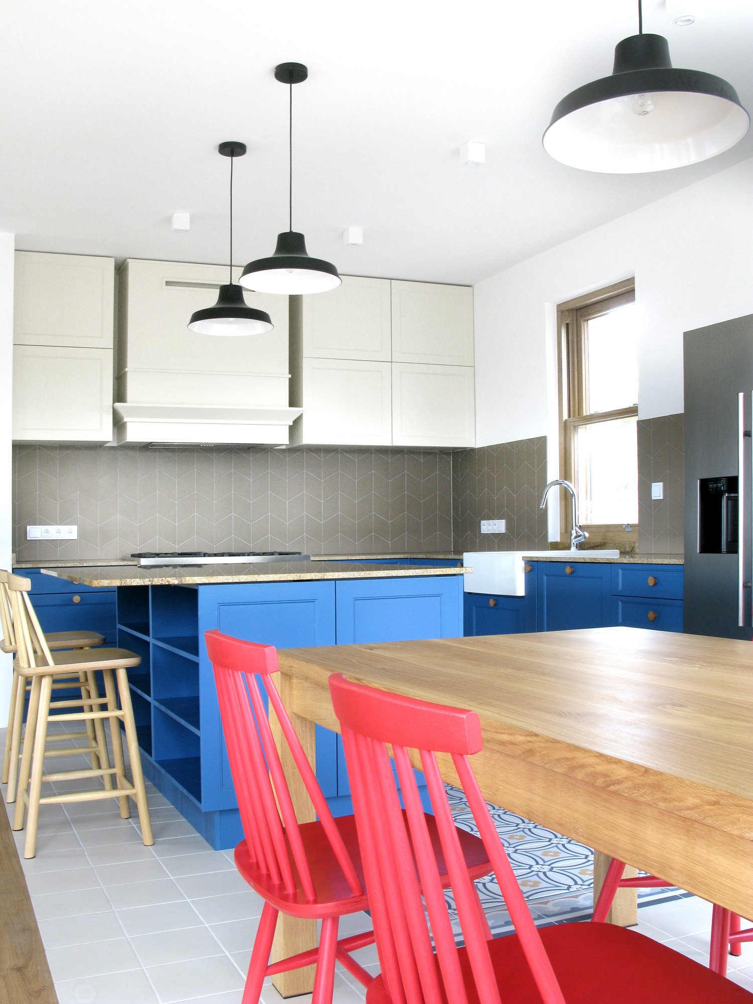Jasna kuchnia z niebieską wyspą, drewnianym stołem i czerwonymi krzesłami w stylu retro, oświetlona trzema wiszącymi lampami.
