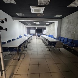Business Horizon Office - Sala konferencyjna z długimi stołami, krzesłami i tablicami. Ciemny sufit z panelami. Podłoga z płytek. Widok w głąb sali, symetryczny układ.
