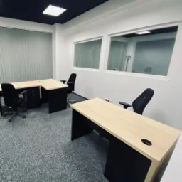 Nowoczesne biuro z dwoma biurkami w kolorze jasnego drewna i czarnymi krzesłami, szara wykładzina dywanowa, okna z widokiem na kolejne biuro, czarny sufit.