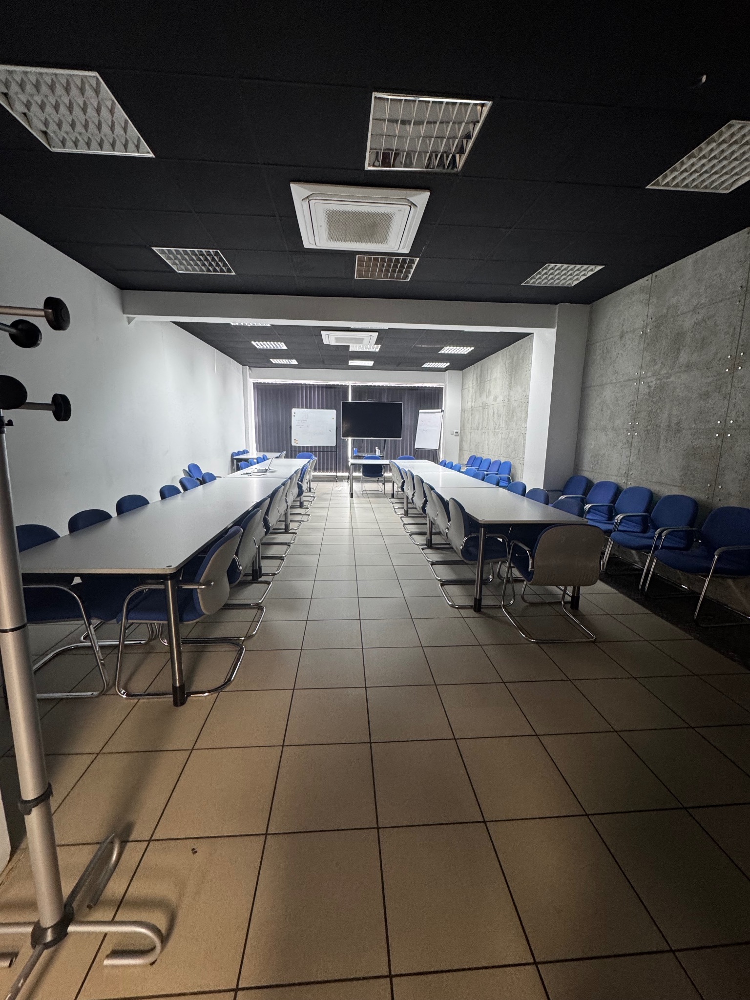 Sala konferencyjna z długimi stołami, krzesłami i tablicami. Ciemny sufit z panelami. Podłoga z płytek. Widok w głąb sali, symetryczny układ.