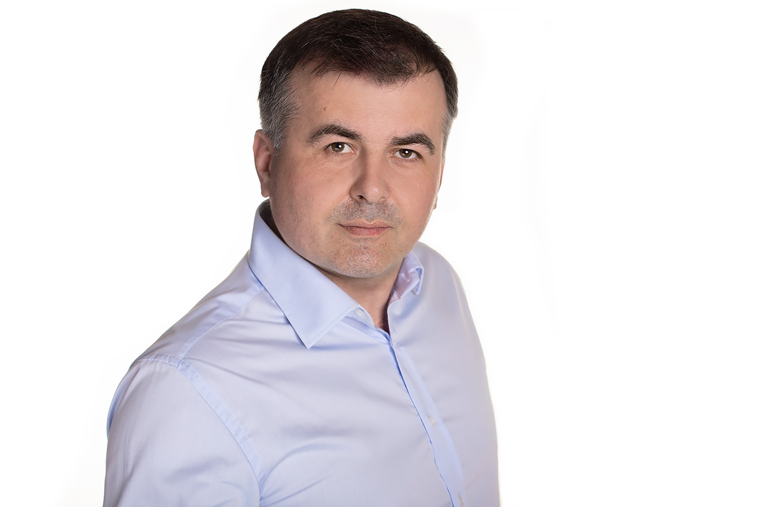 Portret mężczyzny w błękitnej koszuli na białym tle, patrzącego w obiektyw z lekkim uśmiechem.