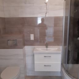 Remonty - Wykończona łazienka z beżowo-białymi płytkami, białą wiszącą szafką z umywalką, toaletą i kabiną prysznicową z zaokrąglonym narożnikiem.