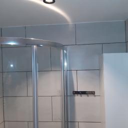 Remonty - Nowoczesna kabina prysznicowa z chromowanymi elementami i szarymi płytkami w łazience, oświetlenie punktowe w suficie.