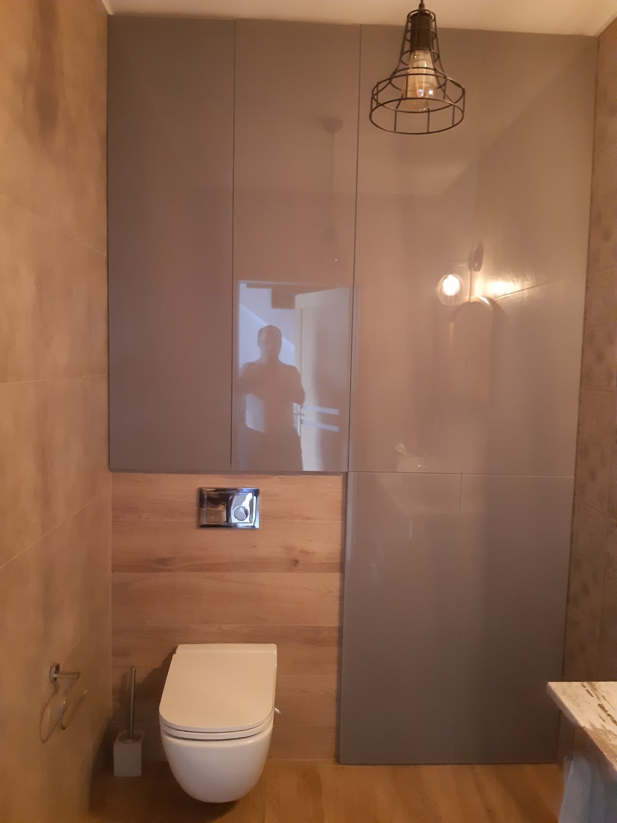 Nowoczesna łazienka z wiszącą toaletą, zabudową meblową w odcieniach szarości i elementami drewna, oświetlona lampą w stylu industrialnym, z odbiciem fotografa w błyszczącej powierzchni szafek.