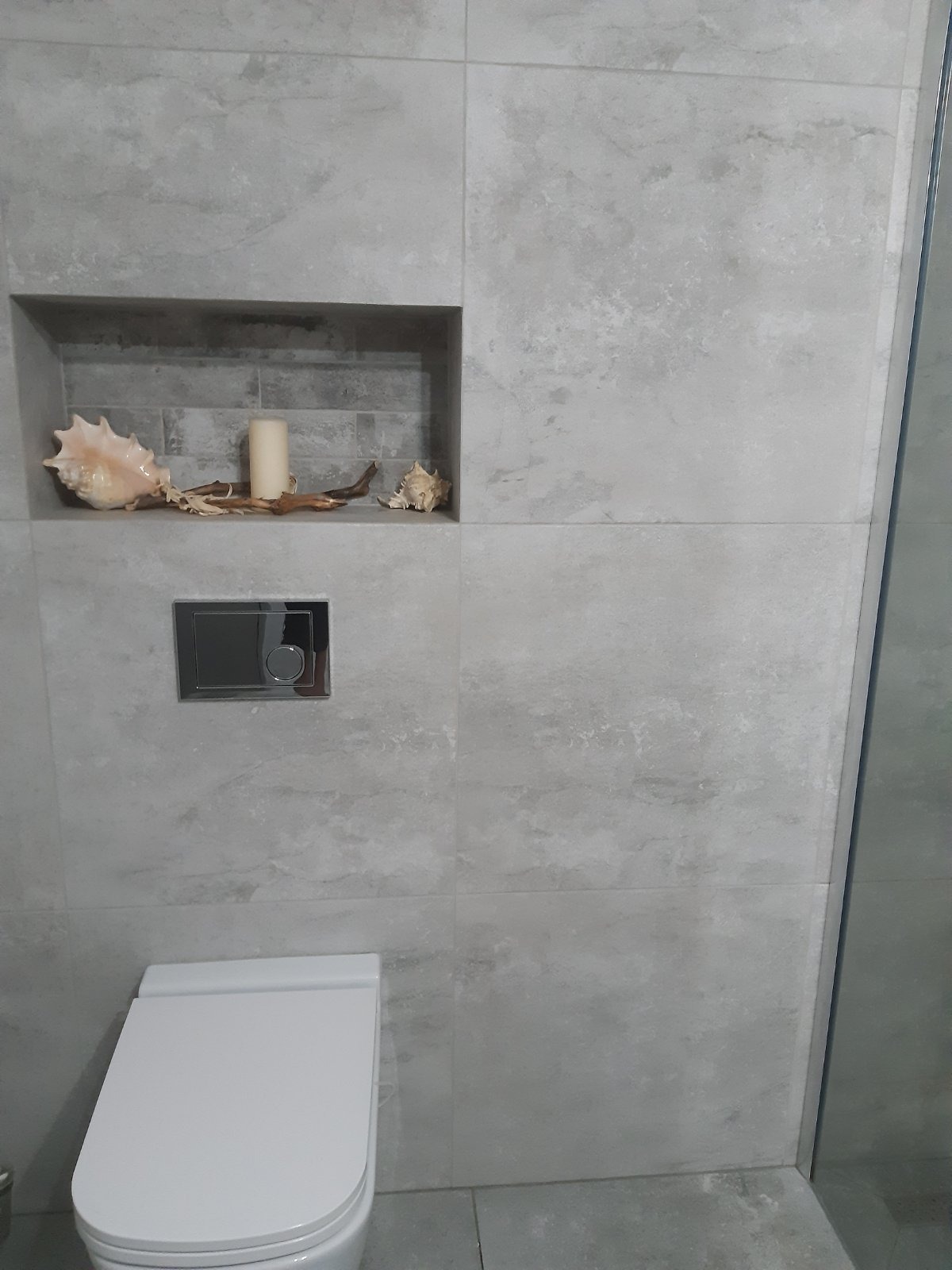 Wykończona łazienka z toaletą podwieszaną i wnęką dekoracyjną z muszlami i świecą, szare płytki imitujące beton.