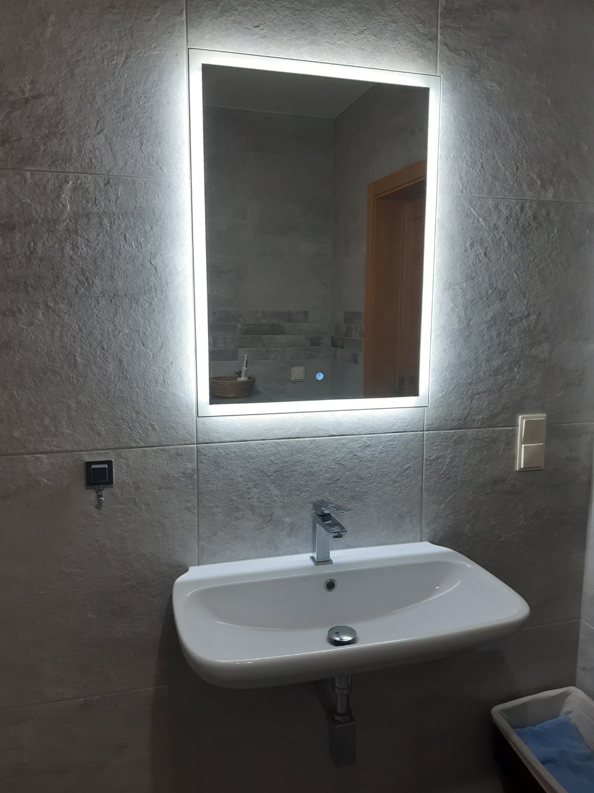 Nowoczesna łazienka z szarymi płytkami imitującymi kamień, podświetlanym lustrem i białą umywalką nablatową z chromowaną baterią.