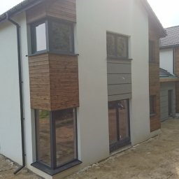 BTS Leszek Szymała - Nowoczesny dom dwukondygnacyjny z elementami drewnianej elewacji i szarą okładziną, widok z zewnątrz na surowym terenie, w tle budowa kolejnego domu.