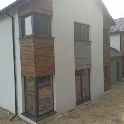 Nowoczesny dom dwukondygnacyjny z elementami drewnianej elewacji i szarą okładziną, widok z zewnątrz na surowym terenie, w tle budowa kolejnego domu.