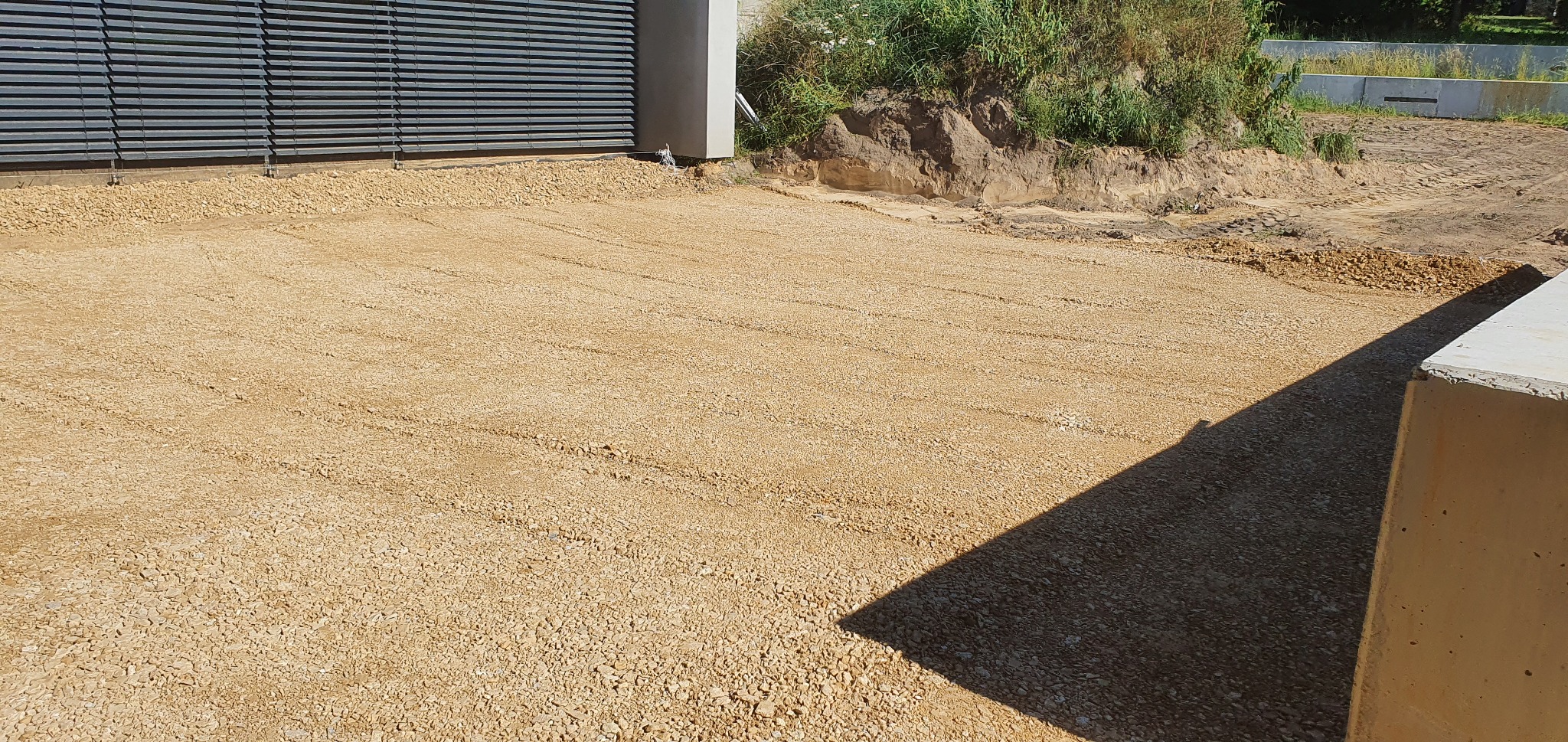 Przygotowany teren pod wylewkę tarasu z widocznymi śladami po wyrównywaniu, sąsiadujący z budynkiem z metalowymi żaluzjami i ziemnym nasypem.