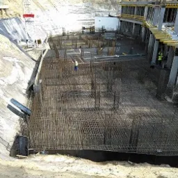 Widok z góry na plac budowy z wykopem pod płytę fundamentową, widoczne zbrojenie w siatce, częściowo zalane betonem, w tle budynek w surowym stanie i żuraw budowlany.