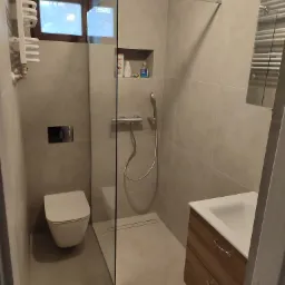 Nowoczesna łazienka z kabiną prysznicową typu walk-in, wiszącą toaletą i szafką z umywalką w odcieniach beżu i brązu. Widoczne detale: odpływ liniowy w podłodze, grzejnik drabinkowy i okno.