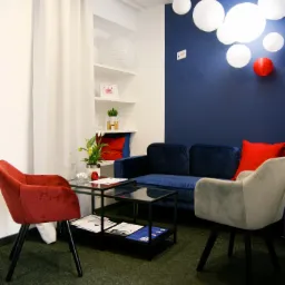 Wnętrze biura nieruchomości w Zielonej Górze z granatową ścianą, białymi lampionami, welurową sofą i krzesłami w odcieniach czerwieni i szarości, stolikiem kawowym z ulotkami informacyjnymi.