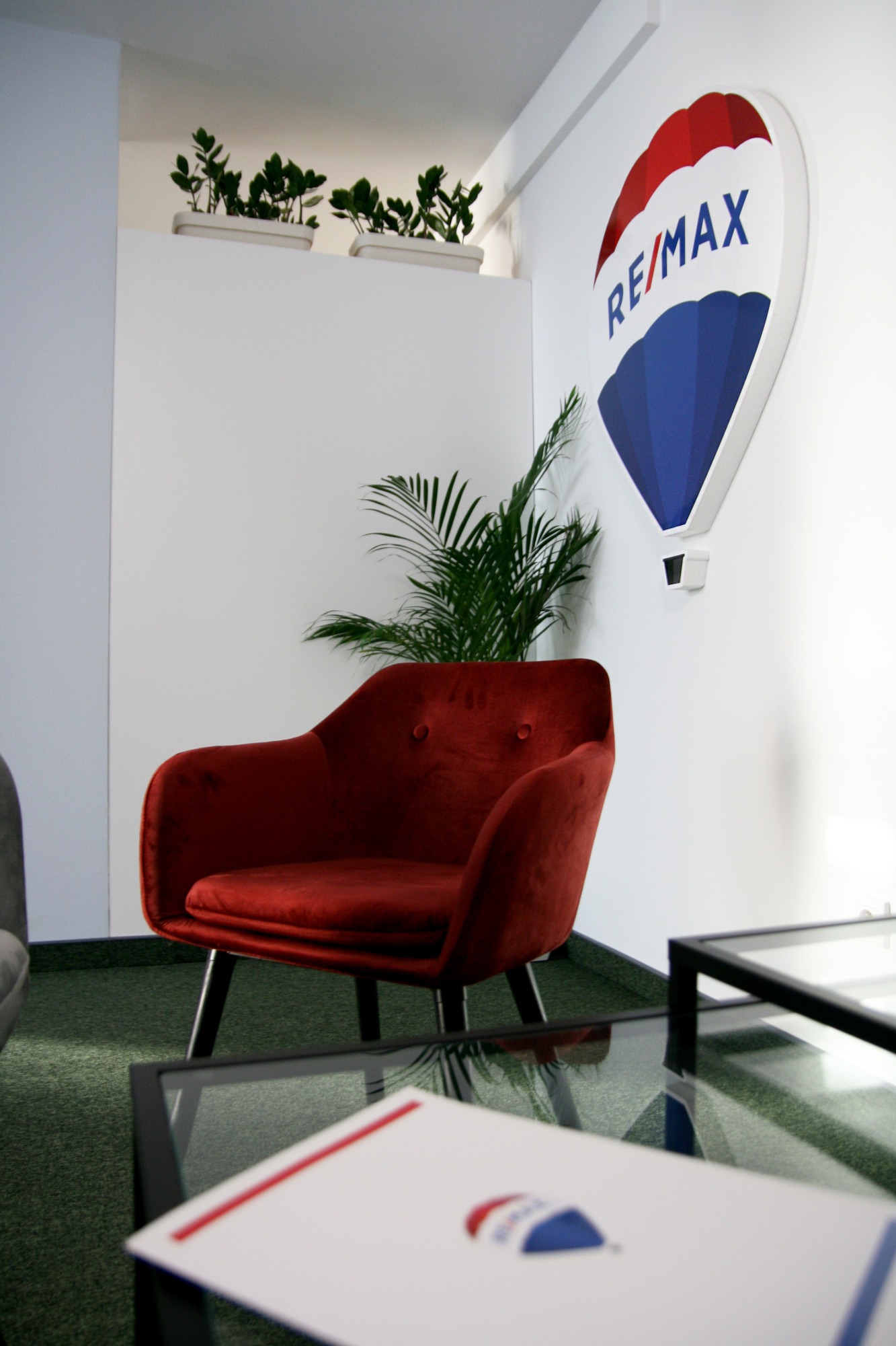 Czerwony fotel w biurze agencji nieruchomości, na ścianie logo RE/MAX w kształcie balonu, na stoliku dokumenty z logo, w tle zielona wykładzina dywanowa i roślina doniczkowa.