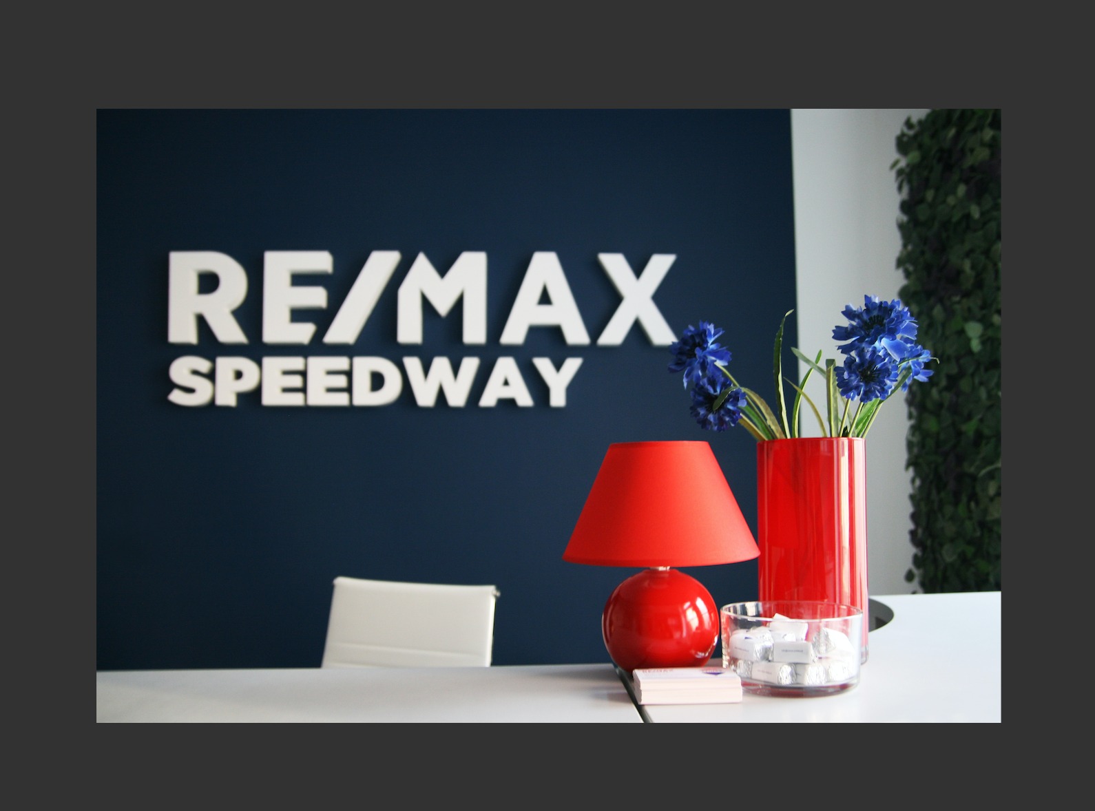 Biuro agencji nieruchomości RE/MAX Speedway z logo na ścianie, czerwona lampka i wazon z niebieskimi kwiatami na białym biurku.