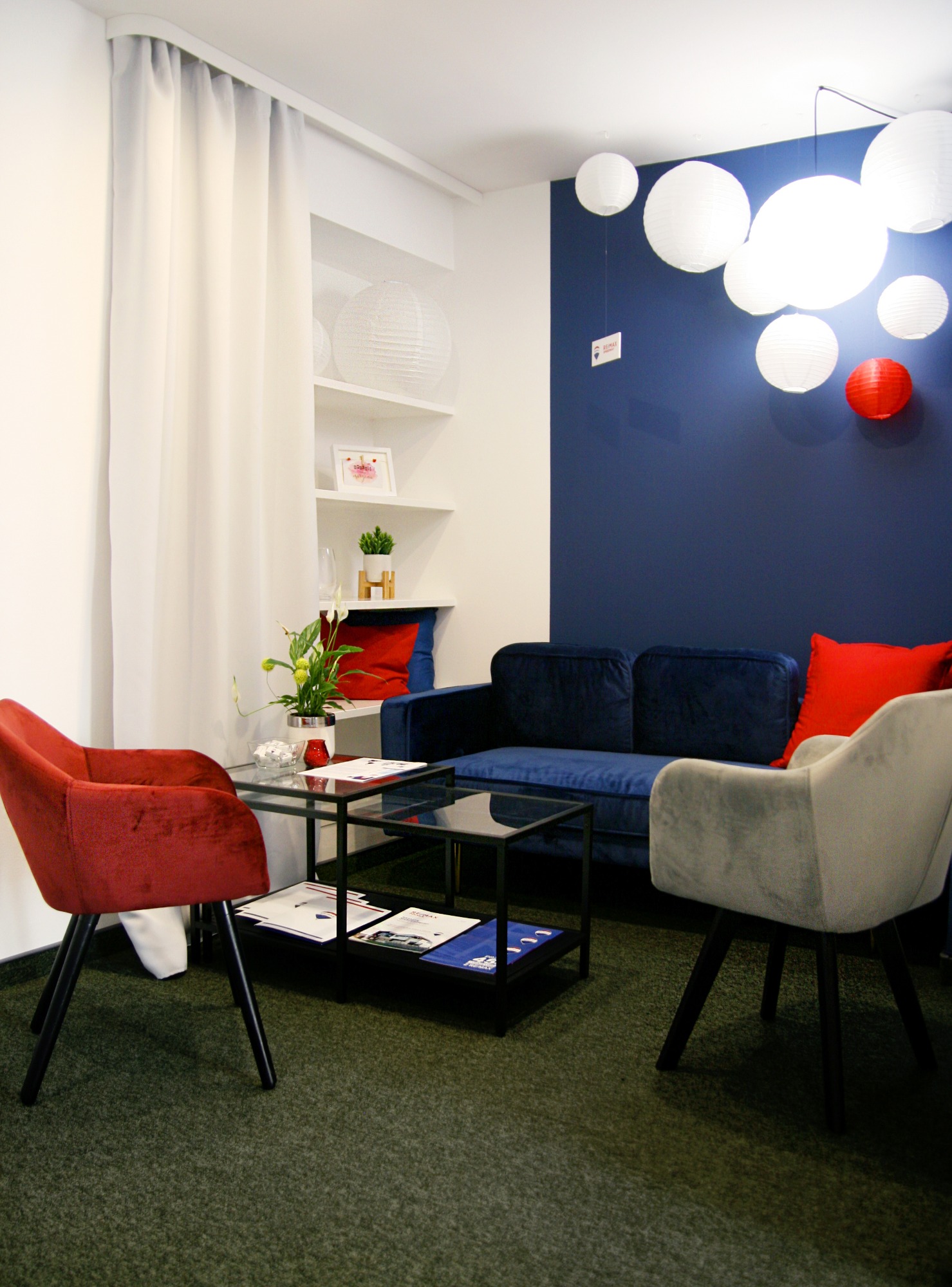 Wnętrze biura nieruchomości w Zielonej Górze z granatową ścianą, białymi lampionami, welurową sofą i krzesłami w odcieniach czerwieni i szarości, stolikiem kawowym z ulotkami informacyjnymi.