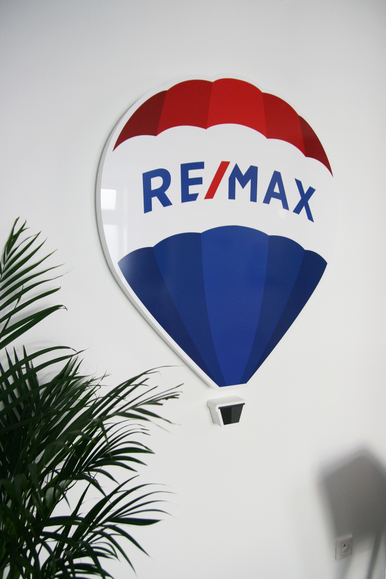Logo RE/MAX w kształcie balonu na białej ścianie, obok zielona roślina doniczkowa.
