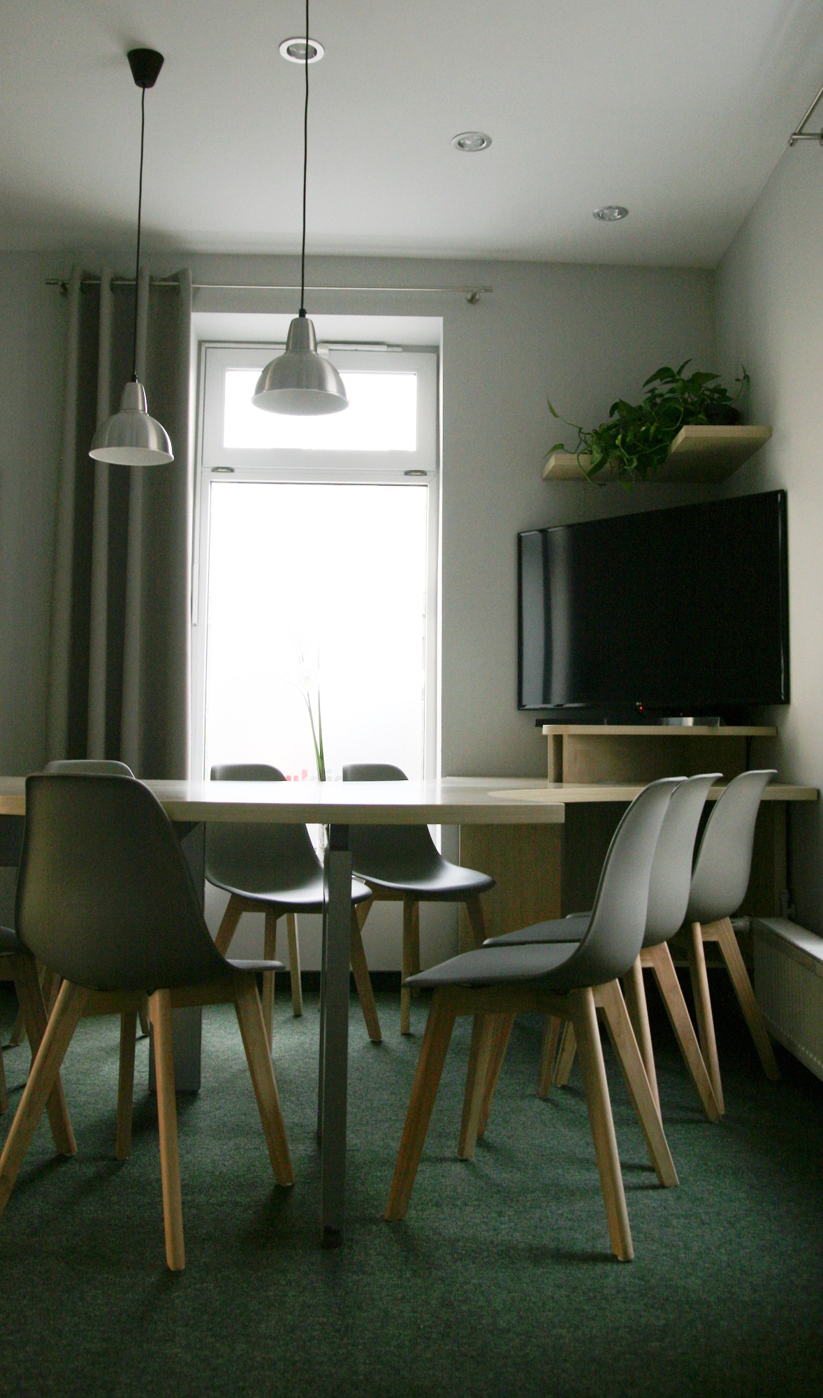 Jasny pokój dzienny z drewnianym stołem, szarymi krzesłami, telewizorem na półce i rośliną doniczkową, oświetlony przez wiszące lampy i światło dzienne z okna.