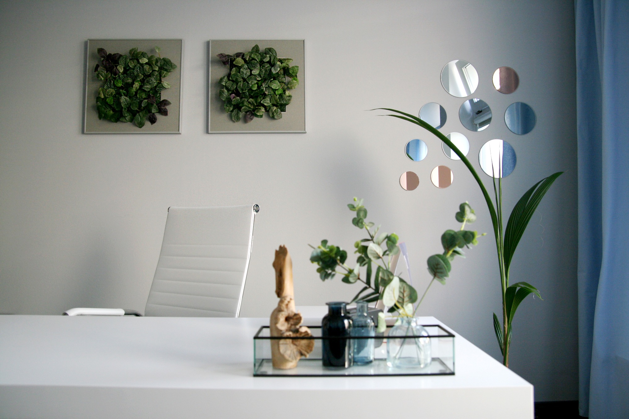 Nowoczesne biuro z białym biurkiem, krzesłem i dekoracjami roślinnymi: ramki z zielenią na ścianie, kompozycja z gałązek w szklanym pojemniku i okrągłe lustra.