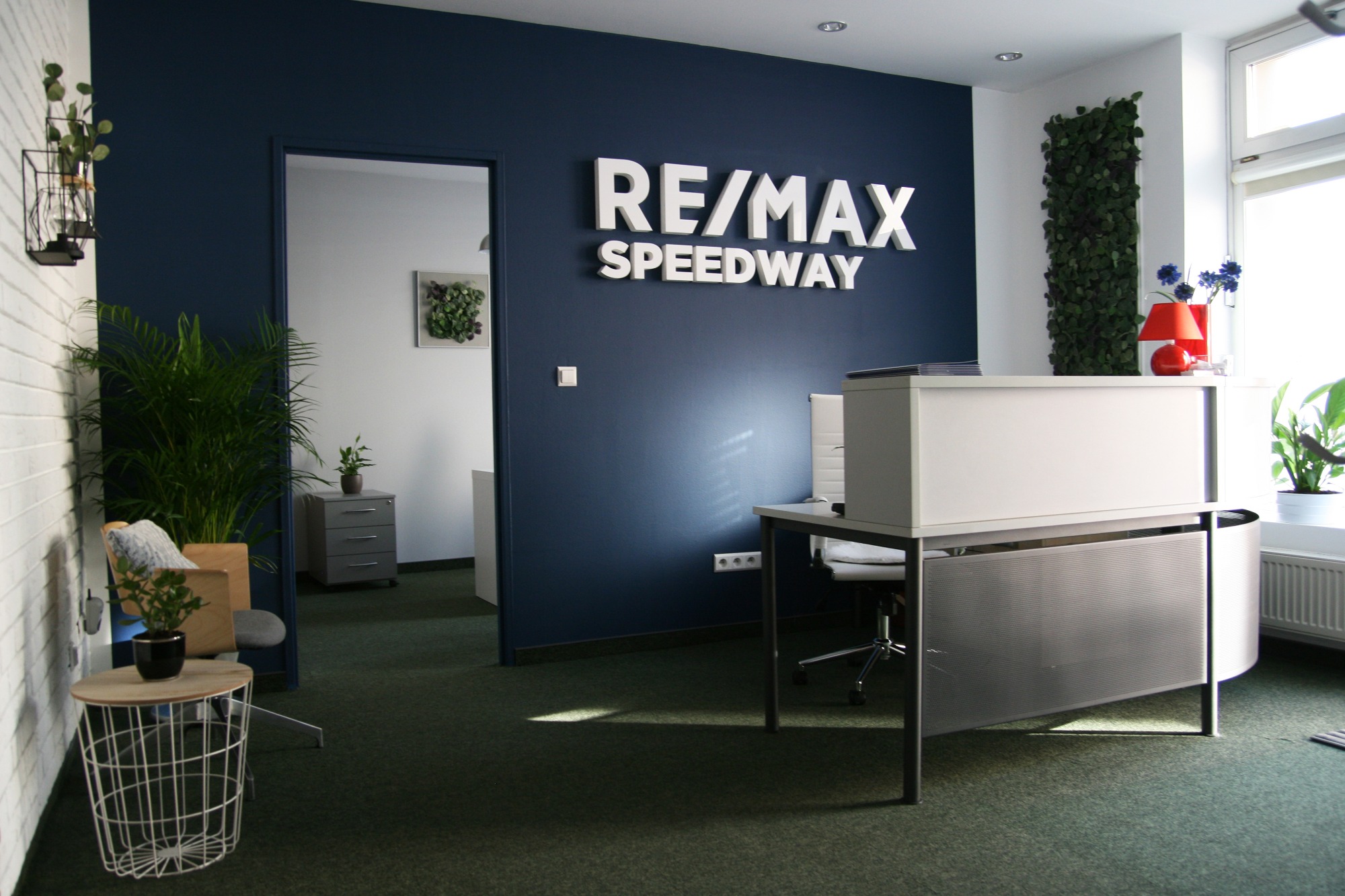 Nowoczesne biuro agencji nieruchomości z recepcją, ciemnoniebieską ścianą z białym logo RE/MAX SPEEDWAY i roślinami doniczkowymi.