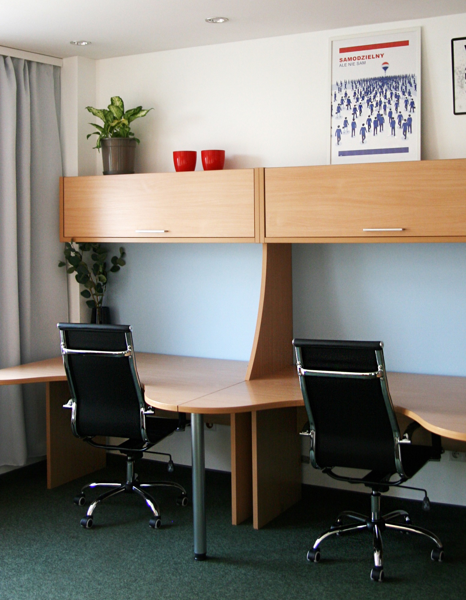 Biuro agencji nieruchomości z dwoma biurkami, czarnymi krzesłami obrotowymi, szafkami wiszącymi i plakatem 'Samodzielny ale nie sam' z logo firmy.