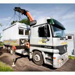 Ciężarówka z dźwigiem HDS podnosi kontener budowlany, widoczny napis 'Transport HDS Kontenery Materiały Budowlane' na pace, w tle drugi kontener.