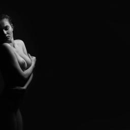 Multiarte - Czarno-biały portret kobiety w akcie, uchwycony w dramatycznym oświetleniu bocznym, z rękami skrzyżowanymi na piersiach i głową pochyloną w dół.