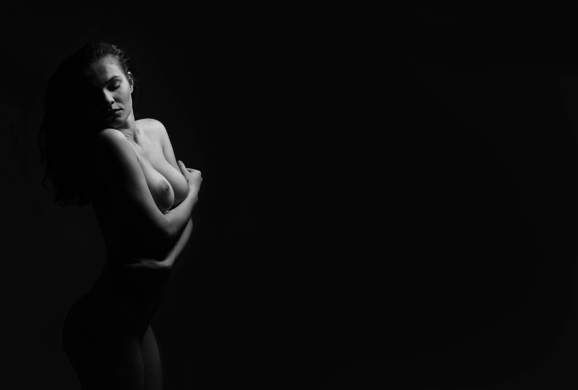 Czarno-biały portret kobiety w akcie, uchwycony w dramatycznym oświetleniu bocznym, z rękami skrzyżowanymi na piersiach i głową pochyloną w dół.