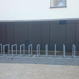 Przedsiębiorstwo Produkcyjno Handlowe MIRAMET Jerzy Kwiatkowski - Montaż stalowej konstrukcji stojaka rowerowego na żwirowym podłożu przed budynkiem z szarymi panelami ściennymi, widoczna drabina i okno na górze.