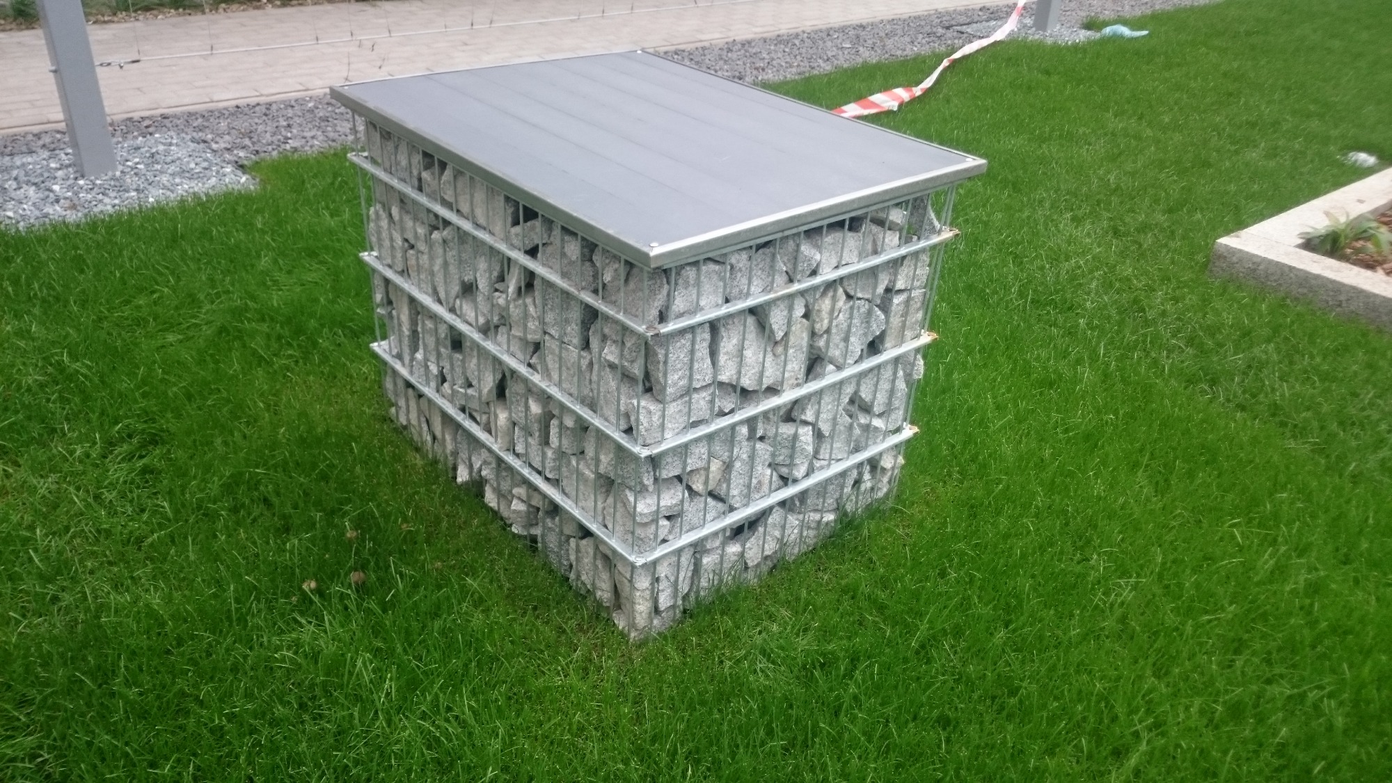 Gabionowa konstrukcja z szarego kamienia, z metalową siatką i szarym blatem, ustawiona na trawniku.