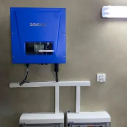 Niebieski inwerter fotowoltaiczny marki BBECO zamontowany na ścianie z widocznymi zabezpieczeniami w białych obudowach i logo Polonus Energy w prawym g&oacute;rnym rogu.