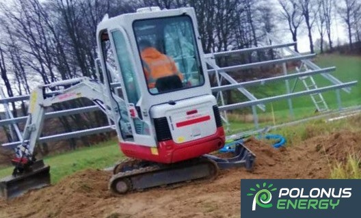 Mini koparka przygotowuje teren pod konstrukcję wsporczą dla paneli słonecznych; operator w pomarańczowej kamizelce w kabinie.