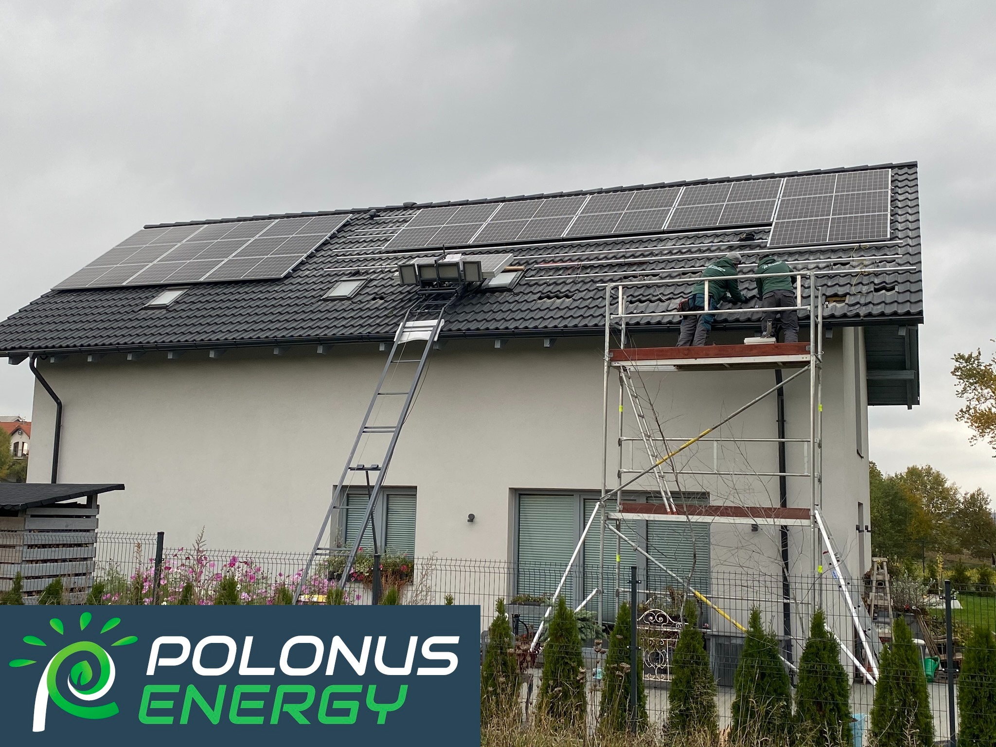 Montaż paneli fotowoltaicznych na dachu domu jednorodzinnego z wykorzystaniem windy dekarskiej i rusztowania, widoczne dwie osoby w uniformach roboczych.