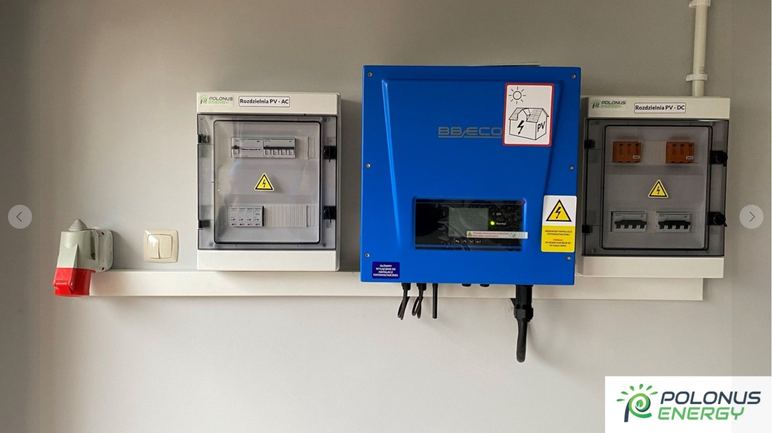 Niebieski inwerter BB ECO zawieszony pomiędzy dwiema białymi rozdzielnicami PV-AC i PV-DC firmy Polonus Energy na białej półce, z widocznym gniazdem siłowym po lewej.