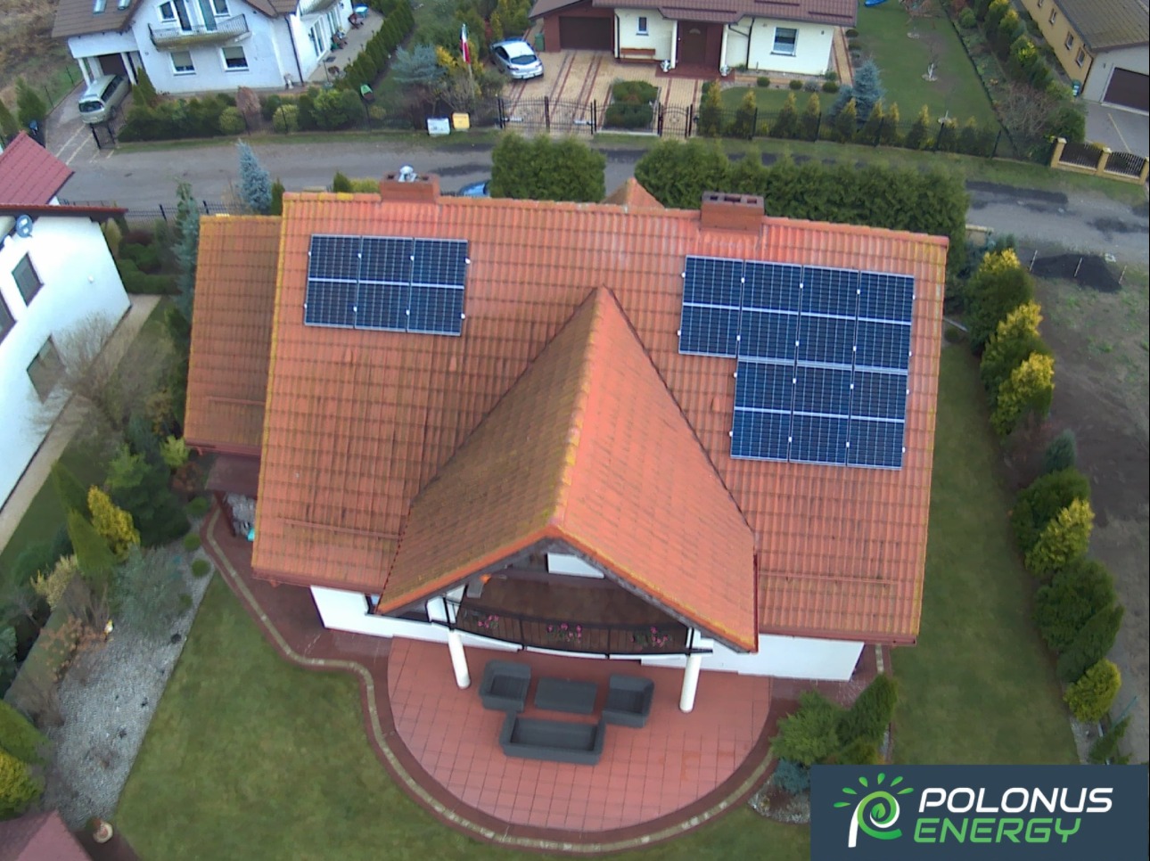 Widok z drona na dom z czerwoną dachówką i panelami słonecznymi zamontowanymi na dachu, w tle inne domy i zieleń.