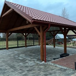 LanDrew - Zadaszona wiata z drewnianą konstrukcją i czerwoną dachówką, widoczne rynny. Posadzka z kostki brukowej. W tle budynek mieszkalny i elementy ogrodu.