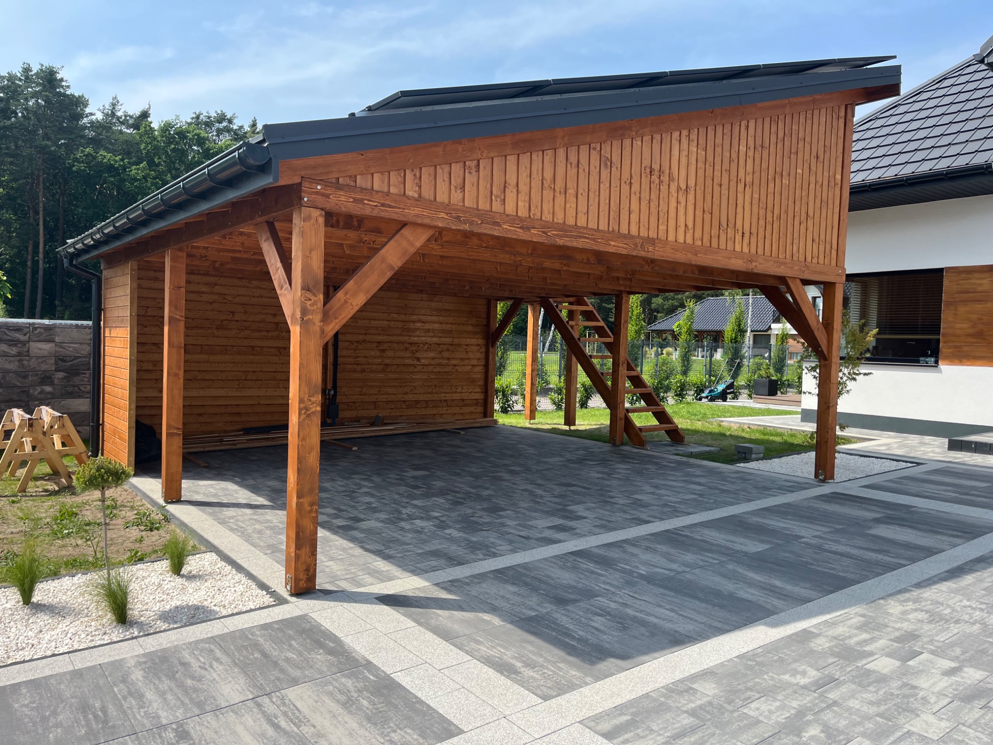 Drewniana wiata garażowa z płaskim dachem i schodami, widoczna z zewnątrz na tle kostki brukowej i zieleni. Wykończenie w ciepłym, brązowym odcieniu drewna.