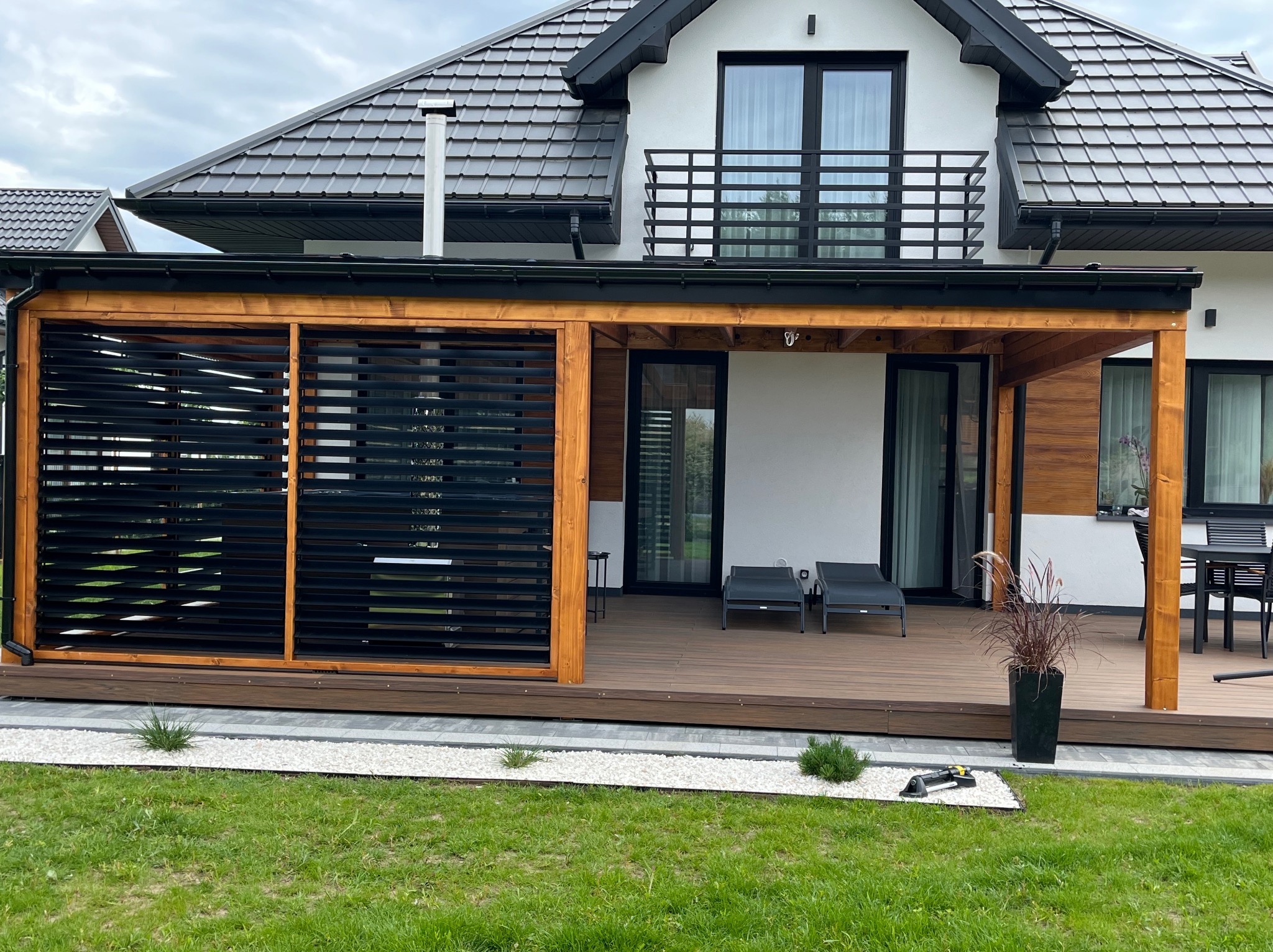 Nowoczesna pergola z drewnianą konstrukcją i czarnymi żaluzjami osłaniającymi taras z leżakami. W tle dom z balkonem i ciemnym dachem.