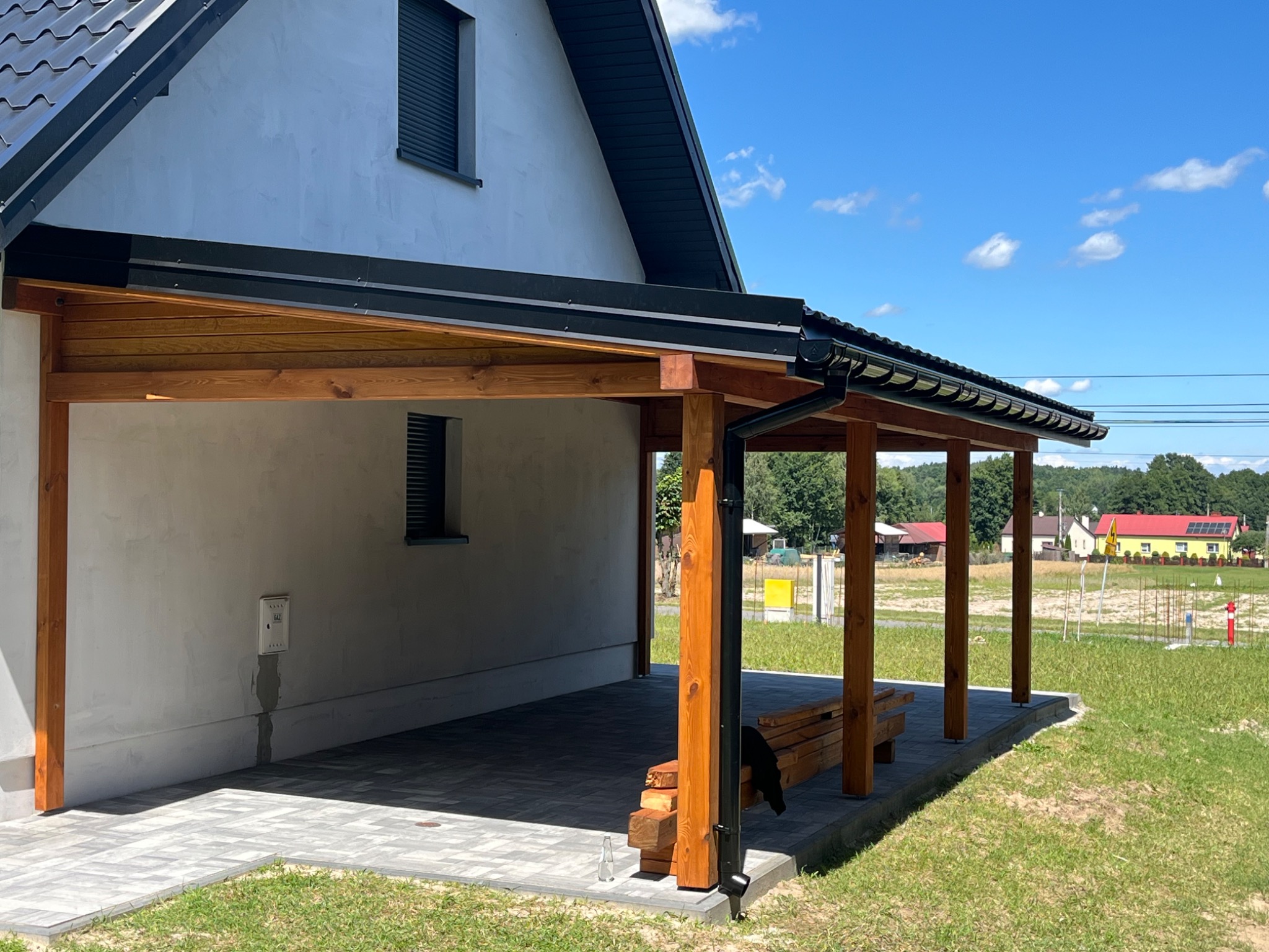 Drewniana wiata przy domu z czarną rynną i kostką brukową. Widok na okolicę w słoneczny dzień. Na pierwszym planie fragment drewnianej ławki.