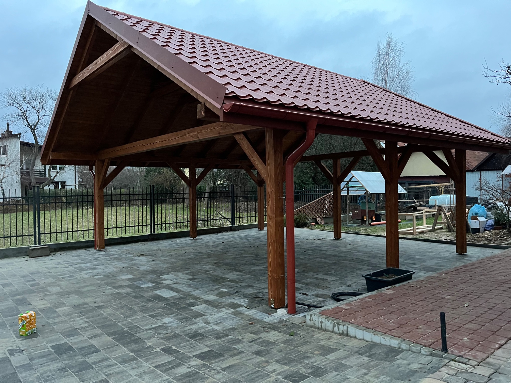 Zadaszona wiata z drewnianą konstrukcją i czerwoną dachówką, widoczne rynny. Posadzka z kostki brukowej. W tle budynek mieszkalny i elementy ogrodu.