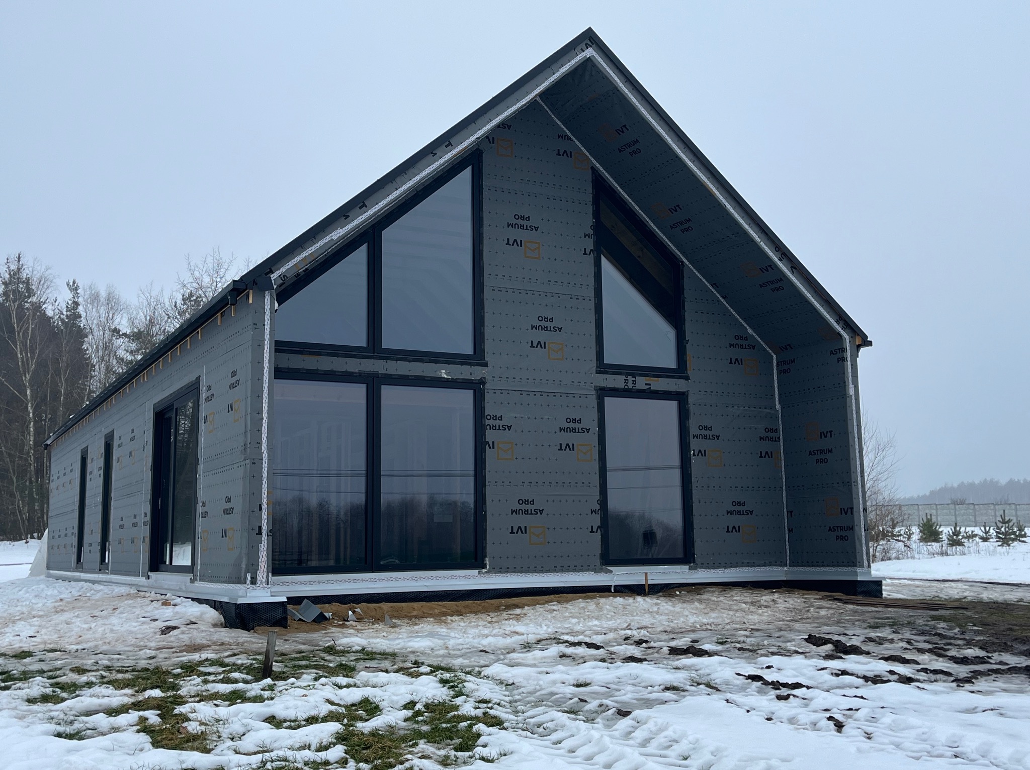 Surowy dom szkieletowy z dużymi oknami, pokryty szarą membraną, w zimowym krajobrazie. Widoczne detale konstrukcyjne i przygotowanie do dalszych prac wykończeniowych.