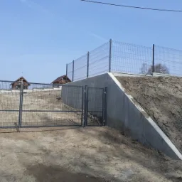 Szara brama panelowa z furtką i ogrodzeniem panelowym na betonowym murze oporowym, zabezpieczającym skarpę ziemną; w tle dwa drewniane domki.