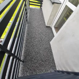 Szary balkon z żywicznej nawierzchni z czarną, metalową balustradą, widok z góry, obok ściana budynku z oknem.