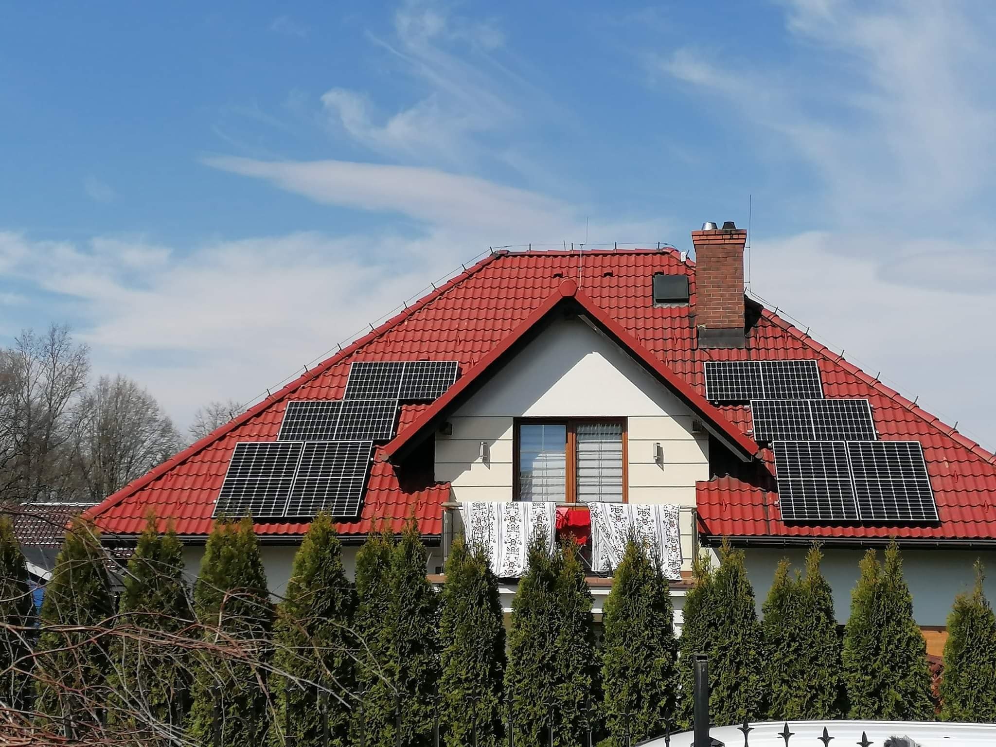 Panele słoneczne na czerwonym dachu domu jednorodzinnego, widoczne pranie na balkonie, błękitne niebo w tle.