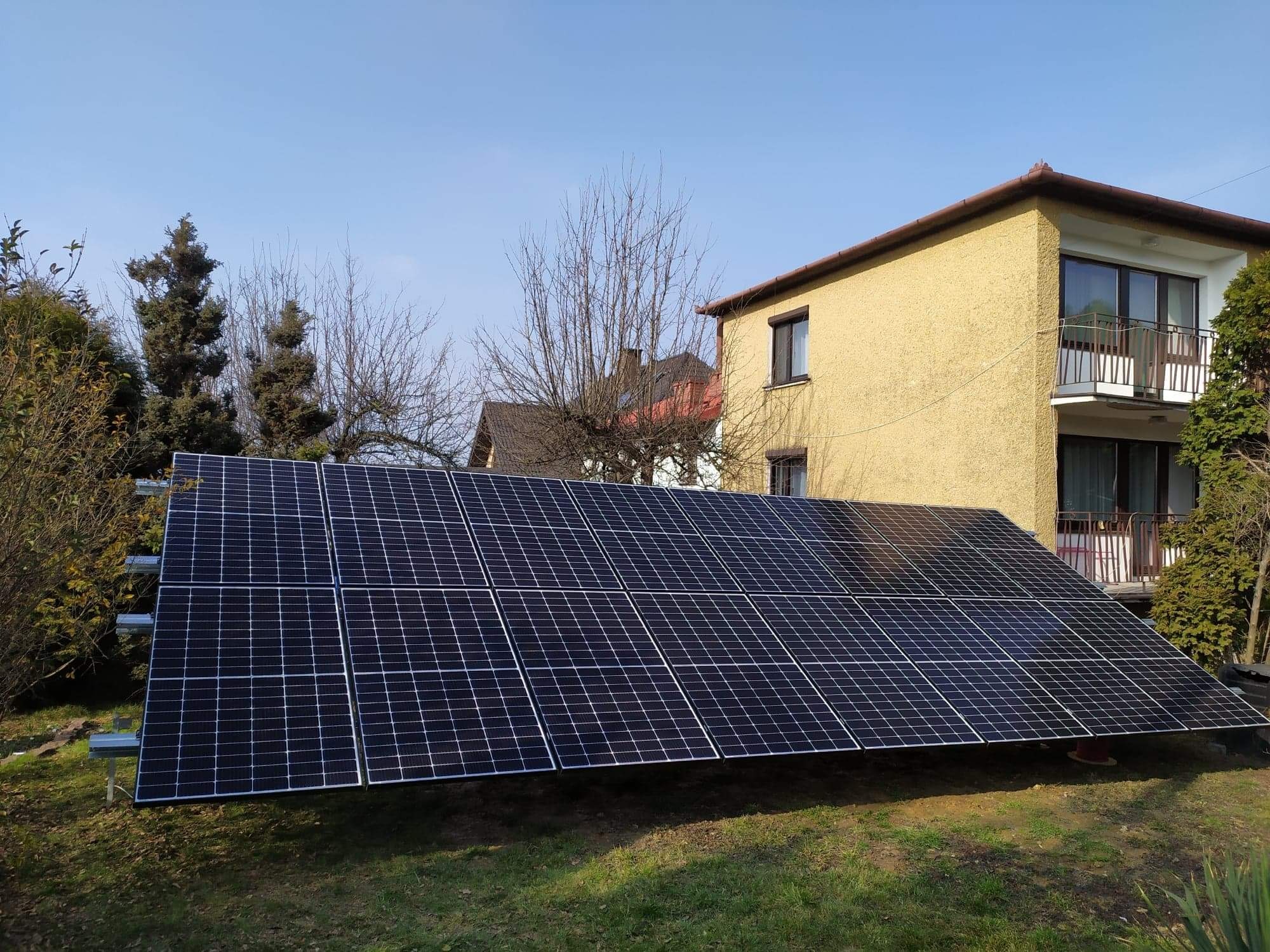 Instalacja paneli słonecznych na trawniku przed dwupiętrowym domem z żółtą elewacją, widoczne drzewa bez liści i krzewy.