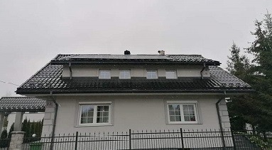 Dom jednorodzinny z zamontowanymi panelami fotowoltaicznymi na dachu pokrytym czarną dachówką, widoczne trzy okna dachowe i komin, pochmurne niebo.