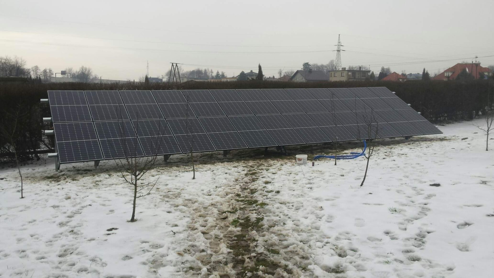 Panel słoneczny w zimowym krajobrazie z odśnieżoną ścieżką, widoczne drzewa bez liści i białe wiadro z niebieskim wężem obok instalacji.