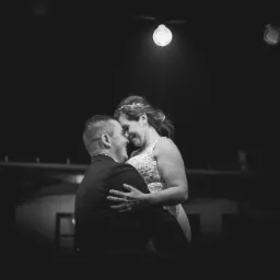 Czarno-białe zdjęcie pary młodej w objęciach, oświetlonej przez żarówkę, z widoczną dekoracją w tle.