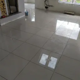 Jasne, błyszczące płytki podłogowe ułożone w dużym pokoju z kominkiem i dużymi oknami, widoczny fragment listwy przypodłogowej i wiadro z wodą.