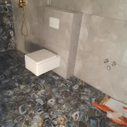 Nowoczesna łazienka z podwieszaną toaletą i podłogą wyłożoną płytkami imitującymi kamienie agatowe, złota bateria natryskowa i szare płytki ścienne.