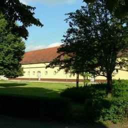 Archiwa Opolskie Sp. z o.o. - Żółty budynek magazynowy z czerwonym dachem, otoczony zielonym trawnikiem i drzewami, widziany zza krzewów.